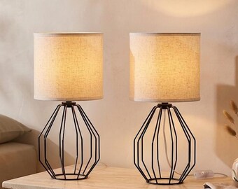 May include: A pair of table lamps featuring black wire cage bases with a geometric, faceted design. Each lamp is topped with a cylindrical, light beige fabric shade that emits a warm glow, creating a soft illumination on the surface.