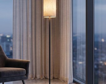 May include: A dark gray upholstered armchair with tapered legs, a slim metallic side table, and a tall floor lamp featuring a thin black pole and a cylindrical beige fabric shade that emits a warm, soft glow in a room with large windows.