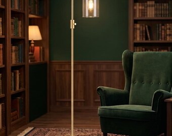 May include: A tall floor lamp with a brushed gold finish, featuring a slender vertical pole, a circular base, and a single cylindrical clear glass shade that houses a glowing light bulb, providing illumination in a room.
