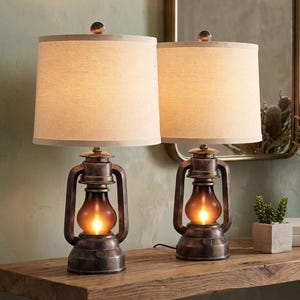 May include: A pair of table lamps featuring a dark bronze-toned metal base designed like a vintage lantern with a glowing amber glass center. Each lamp is topped with a cylindrical off-white fabric shade and a small spherical finial on top.