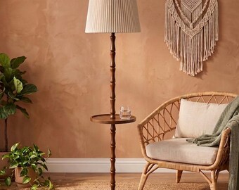 May include: A floor lamp with a dark brown wooden pole, a circular integrated side table, and a pleated off-white drum shade. The lamp stands on a round base and features a clear glass placed on the table surface.