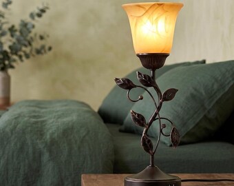 May include: A table lamp with a dark bronze metal base shaped like a vine with leaves. The lamp features a flared, amber-colored glass shade with a marbled pattern. The circular base includes a visible USB port on the side for charging devices.