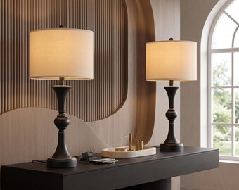 May include: A pair of table lamps featuring matte black metal bases with a curved, pedestal-style silhouette and cylindrical off-white fabric shades. The lamps are positioned on a dark surface, each topped with a small black finial.