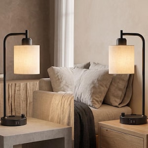 May include: A table lamp featuring a matte black metal frame with a curved arm and a cylindrical off-white fabric shade. The circular base includes integrated USB ports and a rotary control knob for adjusting the light output and power settings.