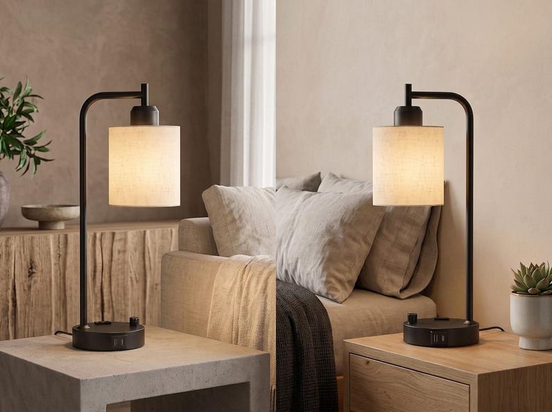 May include: A table lamp featuring a matte black metal frame with a curved arm and a cylindrical off-white fabric shade. The circular base includes integrated USB ports and a rotary control knob for adjusting the light output and power settings.