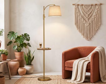 May include: A floor lamp with a brushed gold finish, a circular integrated side table, and a beige fabric drum shade. The lamp features a slender vertical pole, a curved arm holding the shade, and a round base, providing functional lighting.
