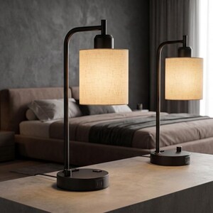 May include: A pair of table lamps featuring matte black metal bases with an L-shaped arm and cylindrical fabric shades in a light beige tone. Each lamp base includes a circular design with integrated power outlets and a small control switch.