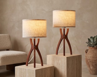 May include: Two table lamps with warm-toned wooden tripod bases and cylindrical fabric shades. The lamps emit a soft, diffused light, highlighting the natural wood grain of the curved legs and the textured weave of the light-colored fabric shades.