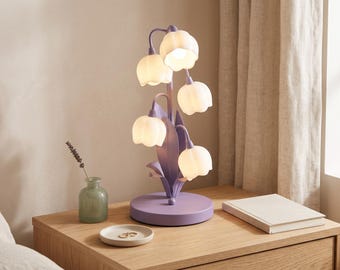 May include: A table lamp featuring a lavender purple metal base and stem shaped like a plant with five white, bell-shaped flower shades that emit a soft glow. The structure includes curved stems and pointed leaves extending from a circular base.