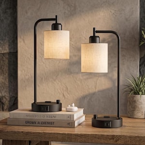 May include: Two table lamps with matte black metal L-shaped arms and cylindrical beige fabric shades. Each lamp base features a built-in electrical outlet and a rotary switch. The lamps are positioned on a wooden surface next to two stacked books.