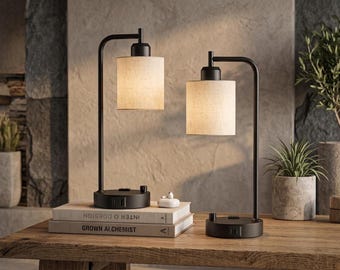 May include: Two table lamps with matte black metal L-shaped arms and cylindrical beige fabric shades. Each lamp base features a built-in electrical outlet and a rotary switch. The lamps are positioned on a wooden surface next to two stacked books.