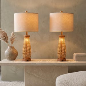 May include: A pair of table lamps featuring cylindrical, light-colored fabric shades and woven, hourglass-shaped rattan bases. The lamps have circular wooden bases and emit a warm, soft glow, illuminating the surface beneath them.