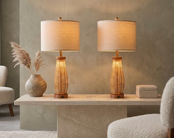 May include: A pair of table lamps featuring cylindrical, light-colored fabric shades and woven, hourglass-shaped rattan bases. The lamps have circular wooden bases and emit a warm, soft glow, illuminating the surface beneath them.