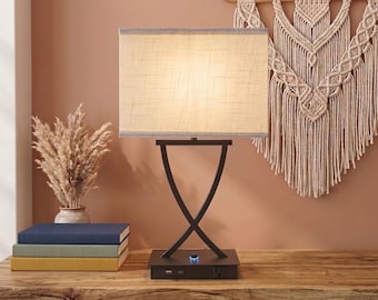 May include: A table lamp with a rectangular beige fabric shade and a dark brown metal base featuring an X-shaped design. The square base includes a power outlet and a USB port, with a small circular power button illuminated by a blue light.