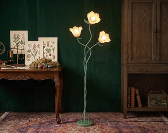 May include: A floor lamp with a green metal base and stem that branches into three intertwined stalks. Each stalk features a white, flower-shaped glass shade containing a light bulb. The lamp stands upright on a circular green metal base.