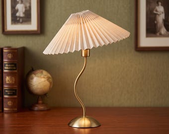 May include: A table lamp featuring a brass-toned, curved, slender stem and a circular base. The lamp is topped with a white, pleated, conical fabric shade that is angled slightly to one side, providing a soft, diffused light from within.