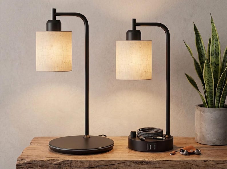 May include: Two table lamps with matte black metal frames and cylindrical off-white fabric shades. The left lamp has a circular base, while the right lamp features a base with integrated USB ports, a rotary switch, and a coiled charging cable.