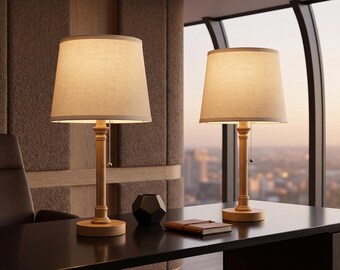 May include: A pair of table lamps featuring light-toned wooden bases with a cylindrical stem and a circular foundation, topped with off-white fabric drum shades and a silver pull chain switch for operation on a dark desk surface.