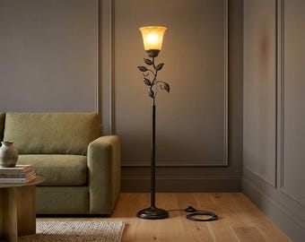 May include: A floor lamp with a dark metal base and a slender stem featuring decorative metal leaves. The lamp is topped with a single, flared, amber-toned glass shade that emits a warm, soft glow, connected to a black power cord with a floor switch.