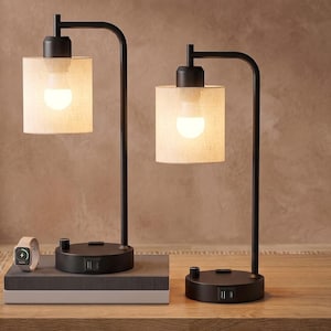 May include: Two black metal table lamps with cylindrical beige fabric shades and illuminated light bulbs. Each lamp base features a rotary switch and integrated USB ports. One lamp sits on a rectangular base, while the other rests on a round base.