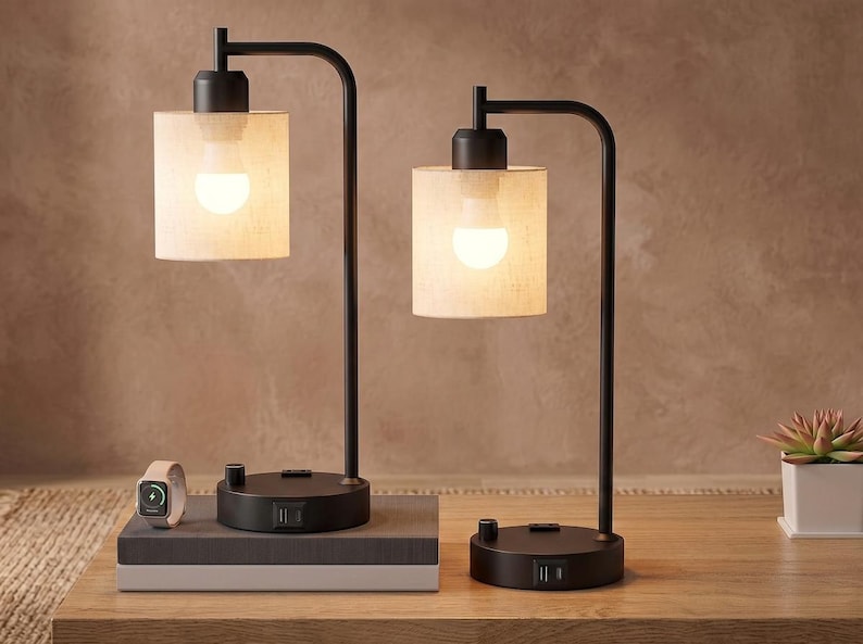 May include: Two black metal table lamps with cylindrical beige fabric shades and illuminated light bulbs. Each lamp base features a rotary switch and integrated USB ports. One lamp sits on a rectangular base, while the other rests on a round base.