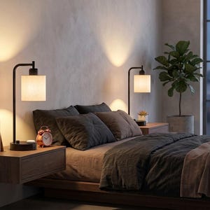 May include: Two black metal table lamps with cylindrical white fabric shades, each featuring a curved arm and a circular base. The lamps are positioned on floating wooden nightstands on either side of a bed with dark gray bedding.