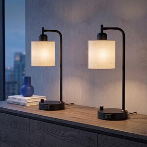 May include: Two table lamps with matte black metal bases and cylindrical off-white fabric shades. Each lamp features a circular base with a rotary switch and integrated USB ports. The lamps emit a warm glow, illuminating the surrounding area.