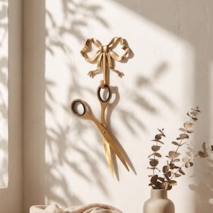 May include: A gold-toned metal wall hook shaped like a decorative bow with a pair of scissors hanging from it. The scissors have dark circular finger loops and long, pointed blades. The wall hook is mounted vertically on a plain white wall surface.