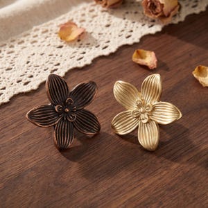 May include: Two flower-shaped napkin rings, one in a dark bronze finish and one in a bright gold finish. Each piece features five elongated petals with vertical line detailing and a small, raised center element with additional petal accents.