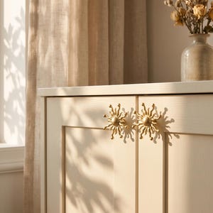 May include: Two gold-toned metal cabinet knobs shaped like suns with radiating rays attached to a light-colored wooden cabinet door. The knobs feature a central circular disc with multiple curved, pointed rays extending outward in a starburst pattern.