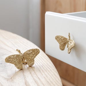 May include: Two gold-coloured metal drawer knobs shaped like butterflies with detailed wing patterns. One knob is attached to a white drawer front, and the other is resting on a light-coloured wooden surface, showcasing the textured wing design.