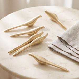 May include: A set of five gold-toned metal bird-shaped objects with smooth, elongated bodies and tapered tails. Each piece features a minimalist, stylized avian form with a matte finish, arranged in a cluster alongside a folded light gray linen napkin.