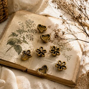 May include: A collection of six small, antique-style brass knobs featuring heart and flower shapes. Three knobs are heart-shaped with textured centres, and three are flower-shaped with five rounded petals, all finished in a muted metallic gold tone.