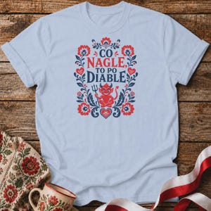 May include: A light blue t-shirt featuring a central graphic with red and dark blue floral folk art patterns. The design includes a small red devil character holding a trident and the text "CO NAGLE, TO PO DIABLE" in a bold, dark blue font.