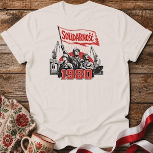 May include: A cream-coloured short-sleeve t-shirt featuring a graphic print of three workers in hard hats standing before a shipyard. A red flag above them reads SOLIDARNOŚĆ, and the red text 1980 is printed prominently at the bottom of the design.