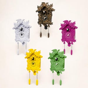May include: Five cuckoo clocks in white, bronze, magenta, yellow, and green. Each clock features a carved leaf and bird motif, a central clock face with black hands, a small bird in a doorway, and two hanging pinecone-shaped weights on chains.