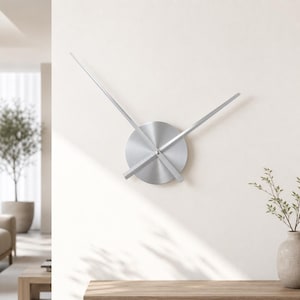 May include: A wall clock featuring a circular, brushed silver metal centre disc with two long, slender silver hands extending outwards. The clock lacks a traditional frame or numerals, presenting a clean, metallic aesthetic mounted on a wall.