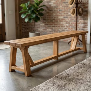 May include: A long, rectangular wooden bench featuring a natural light brown wood grain finish. The bench is constructed with a flat seat surface and sturdy, angled support legs connected by a horizontal base stretcher for structural stability.
