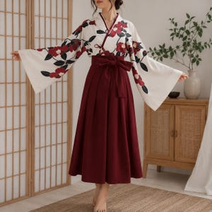 Può includere: Un completo a due pezzi composto da un top bianco in stile kimono con maniche ampie caratterizzato da un motivo floreale rosso e nero, abbinato a una gonna a vita alta color bordeaux scuro con un'ampia fascia in tessuto in vita.