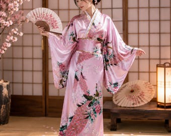 May include: A floor-length light pink satin kimono robe featuring a vibrant peacock and floral print in shades of green, white, and dark pink. The garment has wide, flowing sleeves, a wide matching sash at the waist, and a white collar trim.