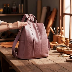 May include: A dusty rose backpack with a textured surface, featuring a vertical stitched seam down the center, a top handle, and adjustable shoulder straps. The bag has a rounded silhouette with side pockets and a main zippered compartment.