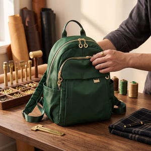Könnte beinhalten: Ein dunkelgrüner Nylon-Rucksack mit goldfarbenen Metallreißverschlüssen mit runden Zippern, einem Tragegriff, verstellbaren Schultergurten und zwei Seitentaschen. Die Vorderseite verfügt über ein Reißverschlussfach und eine kleine goldfarbene Metallplakette.