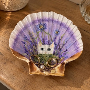 May include: A decorative seashell trinket dish featuring a hand-painted white cat nestled among purple lavender and blue flowers. The interior of the shell is painted in shades of purple and white, and it contains several gold rings and a pearl pendant.