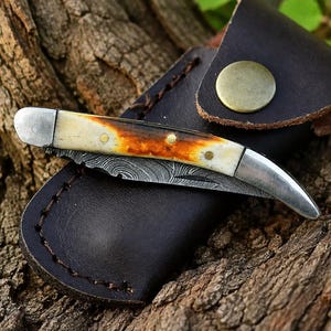 May include: A folding pocket knife with a Damascus steel blade and a cream and amber-colored handle with brass pins, resting on a dark brown leather sheath featuring a brass snap button closure and visible dark brown stitching along the edges.