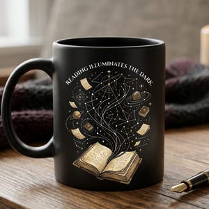 Peut inclure: Un mug en céramique noir mat orné d'un motif doré représentant un livre ouvert d'où s'échappent des lignes célestes, des étoiles, des planètes et des petits livres. Le texte "READING ILLUMINATES THE DARK" est inscrit en arc au-dessus.