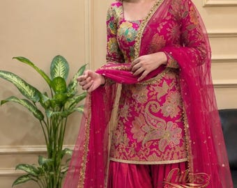 May include: A fuchsia salwar kameez set featuring a long-sleeved tunic with a gold and multi-coloured floral paisley pattern, a square neckline, matching solid fuchsia pleated trousers, and a sheer fuchsia dupatta with gold embroidered trim.
