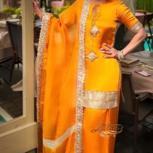 May include: A bright orange salwar kameez set featuring a knee-length tunic with half-length sleeves, a matching pair of loose-fitting trousers, and a long, sheer dupatta. The garment is adorned with intricate gold metallic embroidery along the borders.
