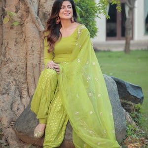 Könnte beinhalten: Eine lindgrüne Tunika mit langen Ärmeln und eine passende Salwar-Hose mit dezentem weißem Punktemuster. Das Outfit enthält einen transparenten lindgrünen Dupatta mit goldbestickten Rändern und verstreuten goldenen Pailletten.
