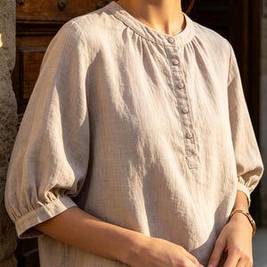 Può includere: Una camicetta beige con scollo rotondo e un'abbottonatura parziale sul davanti con cinque piccoli bottoni. Il capo presenta maniche a tre quarti a sbuffo con polsini arricciati e una finitura in tessuto testurizzato dal look essenziale.