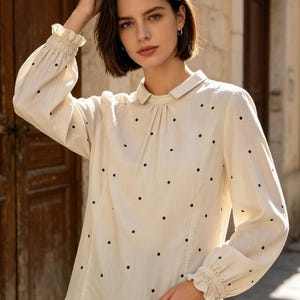 Puede incluir: Una blusa de manga larga color crema con un patrón de lunares negros, cuello clásico y puños fruncidos con bordes de volantes. La camisa tiene un cierre frontal de botones y un corte relajado, confeccionada en un tejido ligero.